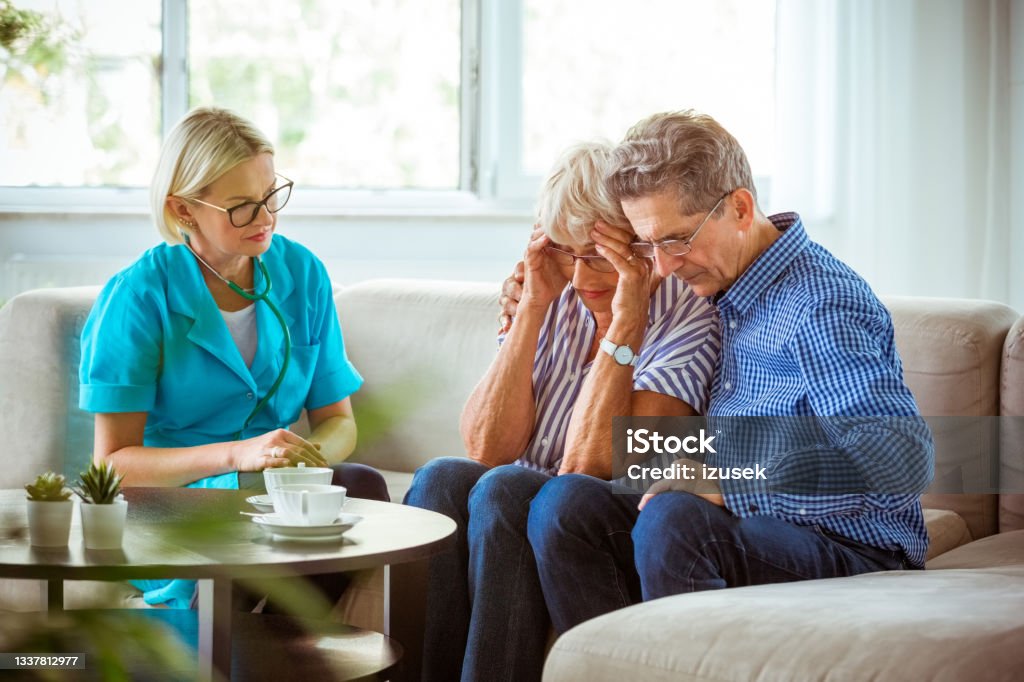 Nurse talking with worried senior couple Worried senior woman feeling unwell, getting bad news. Senior man consoling his wife. They are sitting with nurse in hospital waiting room. Bad News Stock Photo