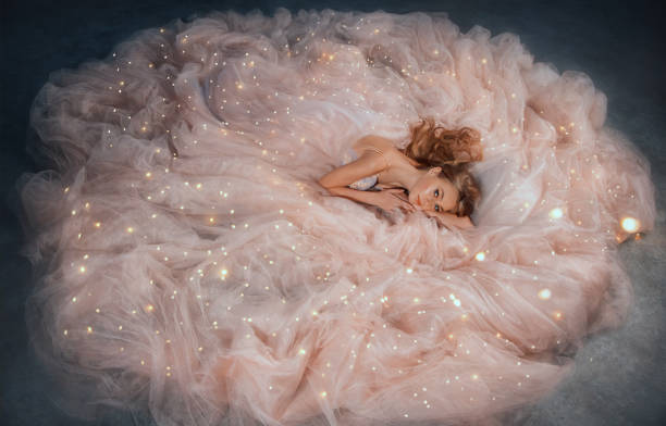 fairy queen fashion model in luxurious shining pink dress posing in studio. princess girl, peach outfit with sequins lies in fabrics of outfit, long skirt gown. fantasy woman sleeping beauty woke up. - evening gown imagens e fotografias de stock