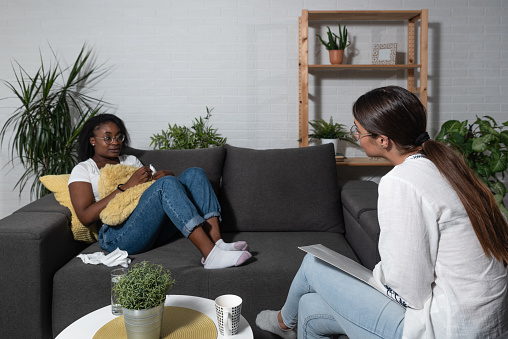 Young black African woman sitting with a psychotherapist in the office has a conversation therapy session trying to solve the problem of depression and anxiety, selective focus