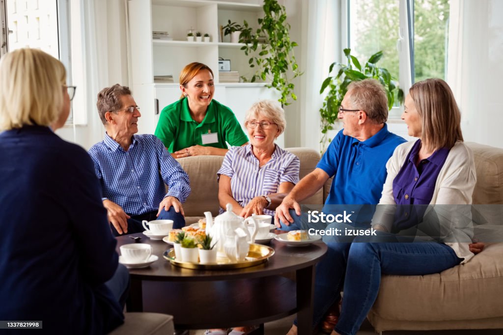 Cheerful senior people in retirement house Happy senior women and man spending time together in nursing home, talking with female social worker, drinking coffee. Nursing Home Stock Photo