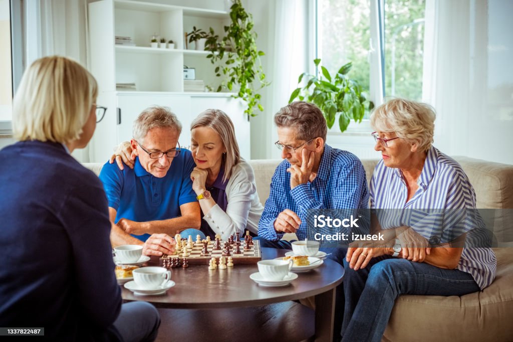 Senior people playing chess in retirement house Senior women and man spending time together in nursing home, playing chess in the living room. Chess Stock Photo