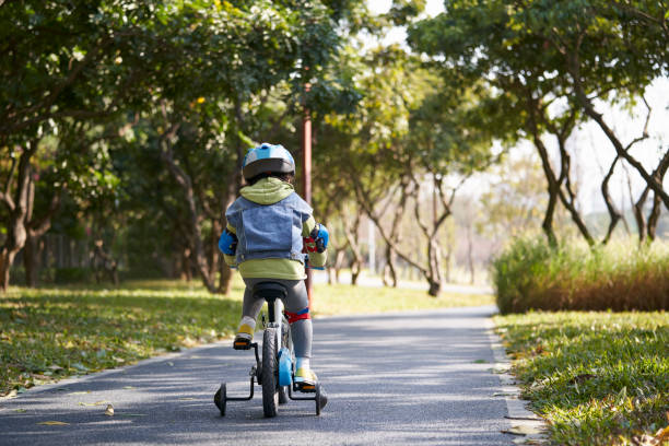 rear view of asian little girl riding bike outdoors in city park - helmet bicycle little girls child imagens e fotografias de stock