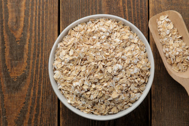 a seco farinha de aveia em uma tigela branca e uma colher de pau. alimento. comida saudável. em uma mesa de madeira marrom. - oat wheat oatmeal cereal plant - fotografias e filmes do acervo