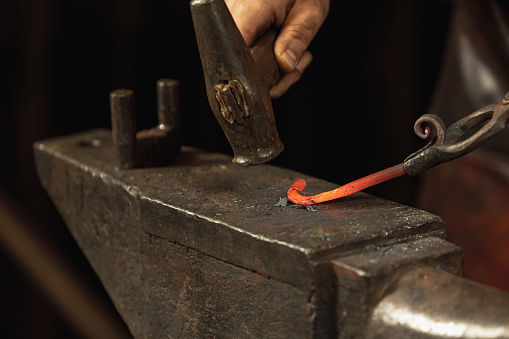 Blacksmith equipment. Close-up working powerful hands of male blacksmith forge an iron product in smithy. Hammer, red hot metal and anvil. Concept of labor, retro professions