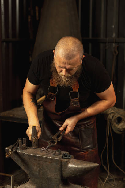 istock Bearded man, blacksmith manually forging the molten metal on the anvil in smithy with spark fireworks. Concept of labor, retro professions, family business 1337829477