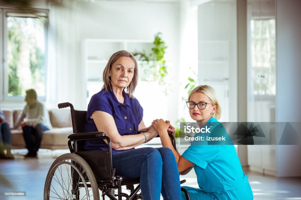 Elderly lady in wheelchair with nurse Home nurse holding hands of worried senior woman in retirement house. Elderly lady sitting in wheelchair, looking at camera. Assisted Living Stock Photo