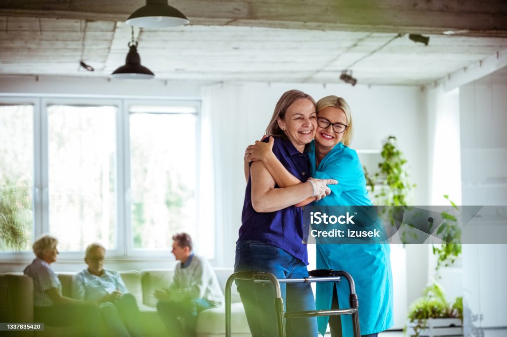 Nurse helping senior woman to use walking frame People in nursing home. Friendly nurse wearing uniform helping elderly lady to use walking frame, they are embracing. 70-79 Years Stock Photo