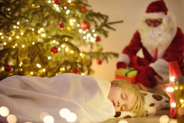 クリスマスイブに木の下でサンタクロースを待っている小さな男の子。子供は眠っています。サンタクロースは贈り物を持ってきました。クリスマスと新年の夜に魔法。 - lying in wait ストックフォトと画像