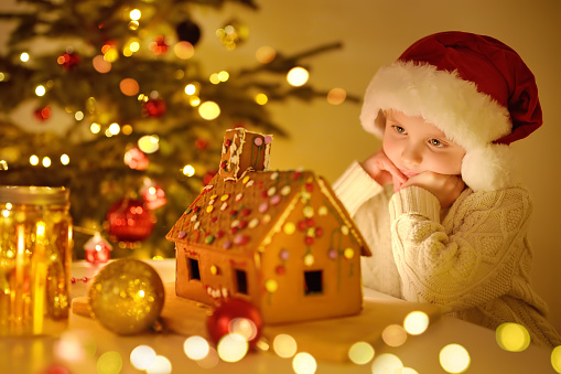Little boy admiring of sweet homemade gingerbread house decorated candies and glazed. Gingerbread house is standing on table near Christmas tree with decorations lights, candles, holidays garlands.