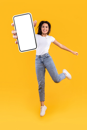 Beautiful young woman recommending new mobile application, vertical collage. Excited lady holding smartphone, showing white blank screen, jumping up over orange yellow studio background, full length