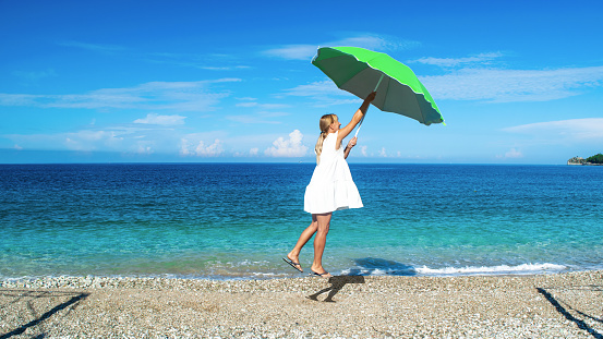 oung girl in a white dress with a green umbrella in her hands is raised by the wind over a pebble beach