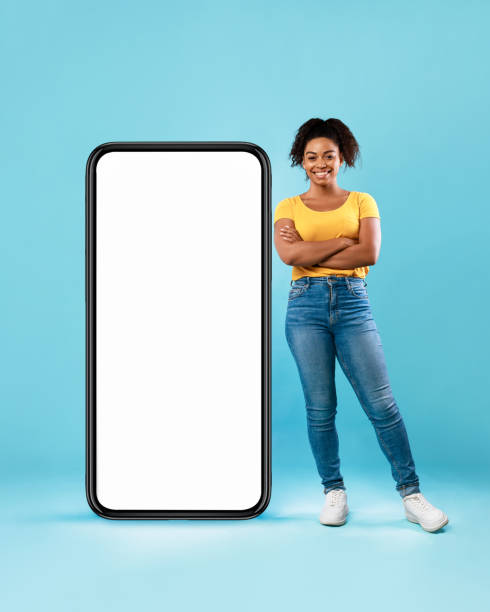 istock Beautiful Afro lady standing near huge smartphone with empty white screen on blue background, mockup. Newest mobile app 1337841392