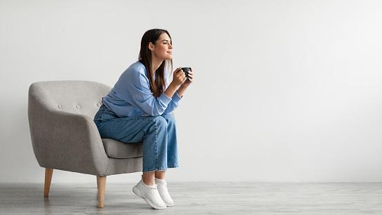 Time for coffee. Charming millennial woman sitting in armchair, enjoying cup of hot drink against white wall