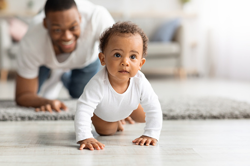 Feliz padre negro mirando a bebé gateando en el suelo de casa photo