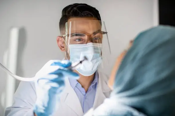 Closeup Shot Of Arab Dentist Doctor In Medical Mask And Face Shield Checking Patient's Teeth, Professional Stomatologist Making Dental Treatment To Muslim Female In Hijab, Selective Focus