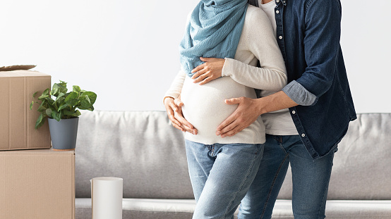 Cropped of pregnant middle-eastern family husband and wife posing at their new house, standing next to sofa and paper boxes with belongings, hugging woman big belly, panorama with copy space