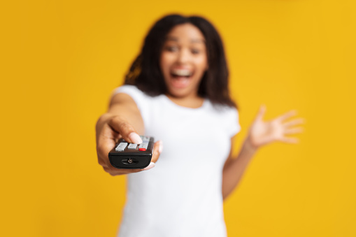 Excited black woman holding remote control in hand, pointing it at camera, watching tv, changing channels, standing at studio over yellow background, selective focus. Relaxation and rest concept