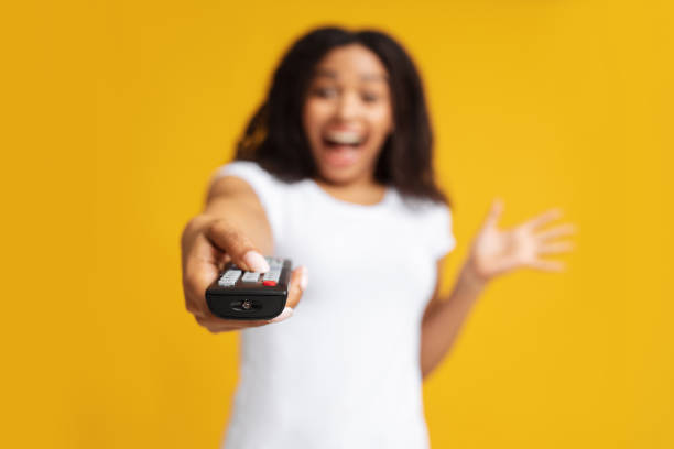 Excited black woman holding remote control in hand, pointing it at camera, watching tv, standing over yellow background Excited black woman holding remote control in hand, pointing it at camera, watching tv, changing channels, standing at studio over yellow background, selective focus. Relaxation and rest concept control point stock pictures, royalty-free photos & images