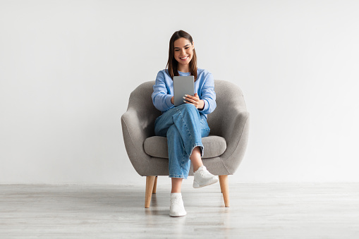 People, technology and communication concept. Portrait of smiling young woman with digital tablet sitting in armchair, working online against white studio wall, full length