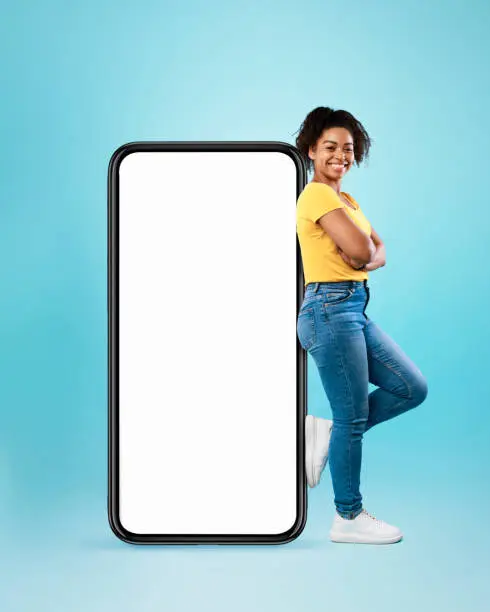 Photo of Black woman leaning on cellphone with space for mobile app on white screen, blue background. Smartphone display mockup