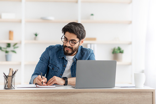 Positive arab male manager working from home during COVID-19 pandemic, using modern laptop and taking notes, sitting at workdesk in living room interior, copy space