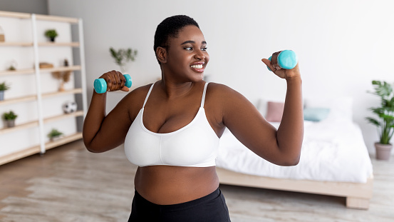 Plus size young black lady exercising with dumbbells at home during covid lockdown, banner design. Happy overweight Afro woman strengthening arm muscles, having weight loss training indoors