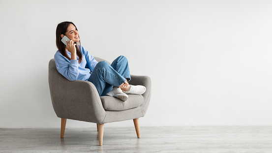 Friendly millennial woman speaking on mobile phone, sitting in armchair, enjoyng conversation with friend against white wall, banner with empty space. Modern communication concept