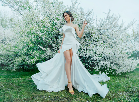 Fantasy woman in long white elegant fashion long dress walks in green spring blossom cherry garden. Happy cheerful girl princess bride. Skirt fabric flies flowing waving in wind motion. Sexy bare legs