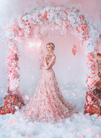 Fantasy spring happy woman in blooming long dress. Fabric skirt in flowers. Creative arch with red, pink white roses. Petals in the snow. Fairy princess girl. Blond hair. Fashion model queen of glamor