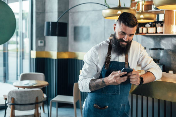 Happy Barista Using Mobile Phone in a Cafe Cheerful smiling waiter with a beard leaning on the bar counter and typing text message on his smartphone while working at the coffee shop. waiter stock pictures, royalty-free photos & images