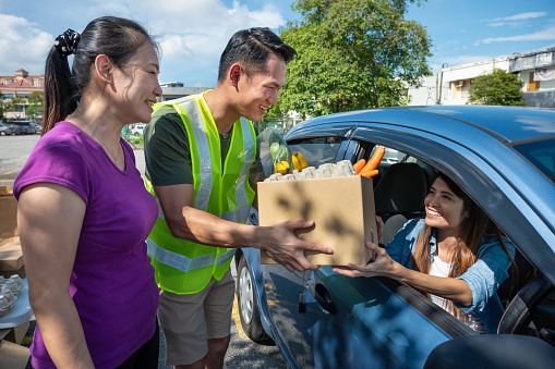 Young volunteers cheerfully hands a paper bag full of groceries for car driver through the car window.