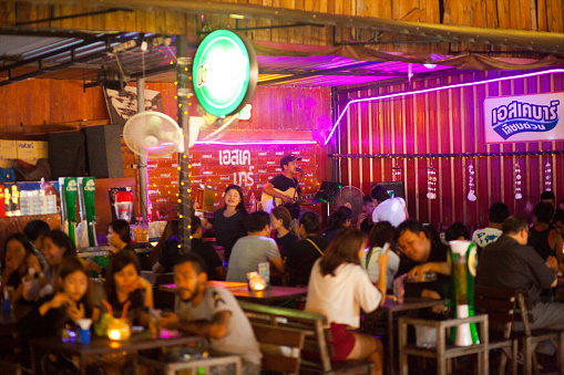 Young adult thai people in bar on night market Liab Duan in Bangkok Ramintra. people are sitting in groups at tables. In background one male singer and musician is playing guitar in corner of bar