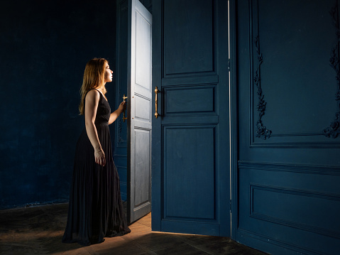 young woman in black dress opens a door from which light is pouring. The dark room is illuminated by mysterious light from behind the door. concept of discovering secret knowledge and opportunities