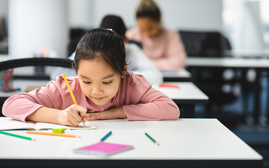 Tuition Concept. Portrait of concentrated small asian girl sitting at table in classroom, writing or drawing in notebook, doing homework or taking notes during lesson, selective focus, free copy space