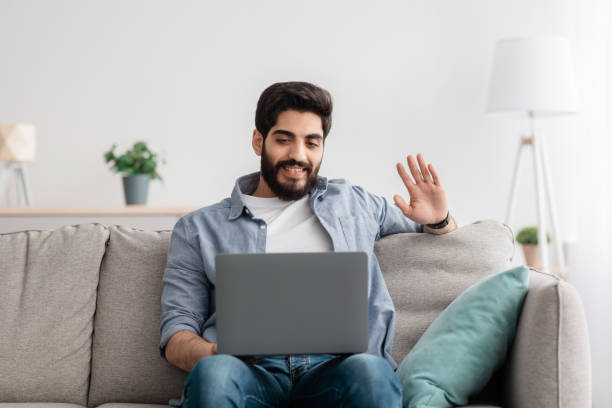 istock Online communication. Cheerful arab guy talking at laptop camera, relaxing on sofa at home interior, free space 1337890486