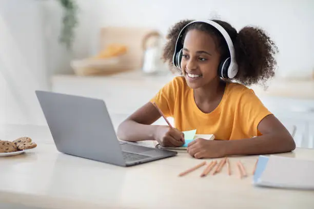 Photo of Cheerful black girl studying online, using laptop and headset, drawing