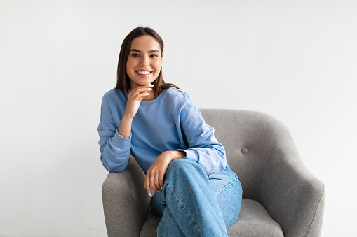 Happy lady. Smiling young friendly Caucasian woman wearing casual clothes, looking at camera, sitting in armchair, posing on white studio background. Real people portraits concept