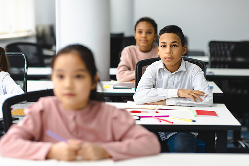 First Day At School. Portrait of focused diligent boy sitting with folded arms at table with group of classmates in blurred background, listening to teacher, selective focus. Back To Study