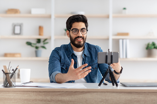 Arab male blogger recording video at home with smartphone on tripod, middle-eastern man making content for his blog, sitting at home office and gesturing at phone camera, copy space
