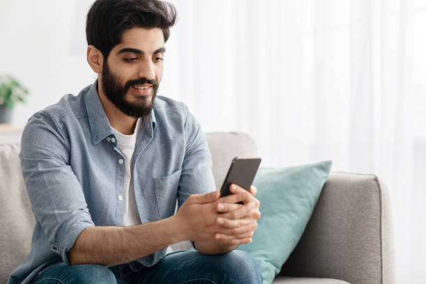istock Mobile communication. Happy arab man reading messages on smartphone at home while relaxing on couch, free space 1337891515