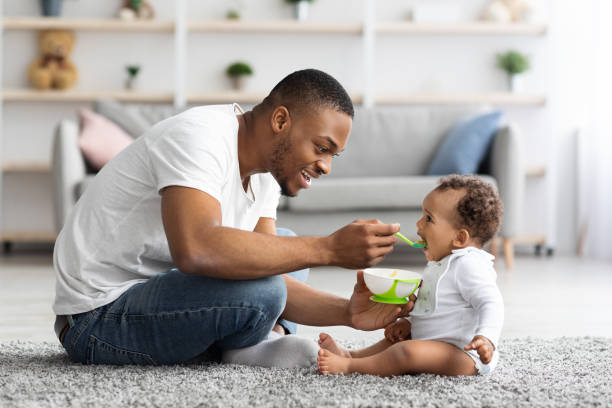 Father's Care. Loving Black Dad Feeding His Cute Baby Son At Home Father's Care. Loving Black Dad Feeding His Cute Baby Son From Spoon At Home, Young African American Daddy Giving Healthy Food To His Little Toddler Child While They Relaxing Together In Living Room infant feeding stock pictures, royalty-free photos & images