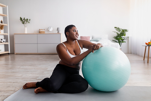 Curvy black lady sitting near fitness ball, exercising on sports mat at home, full length. Young overweight Afro woman doing domestic workout, training indoors during covid quarantine