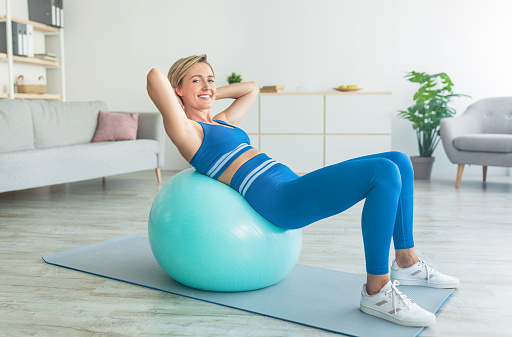 Domestic Train Concept. Smiling sporty middle-aged woman doing abs exercises at home, leaning on swiss fitness ball, looking at camera. Positive lady working out in living room with sports equipment