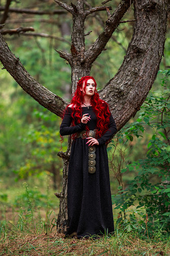 Courageous young lady with long red hair in the image of a fabulous historical character of a witch and a priestess in a mystical forest