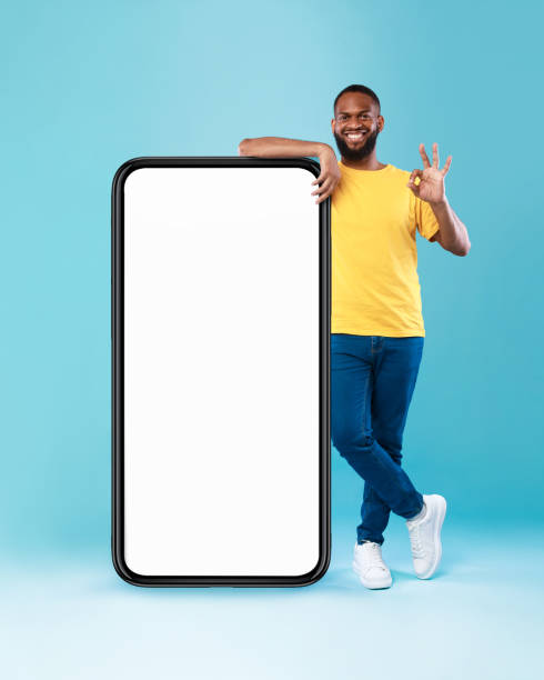 istock Cool black guy showing okay gesture next to huge smartphone on blue background, mockup for mobile app design on screen 1337891766
