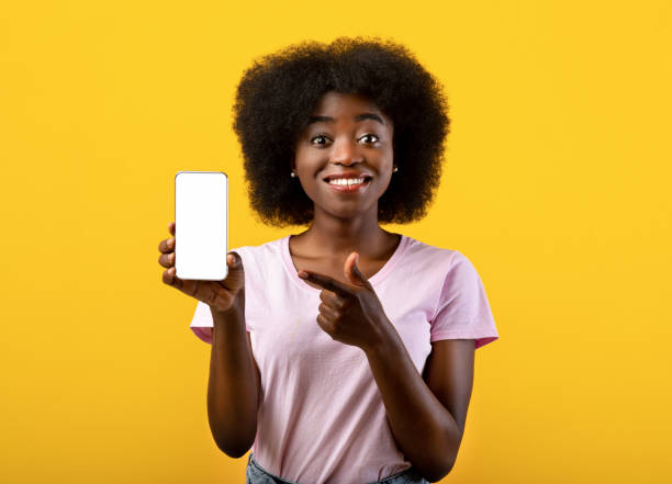 istock Cheerful african american lady pointing on smartphone with white empty screen in hand, standing over yellow background 1337891785