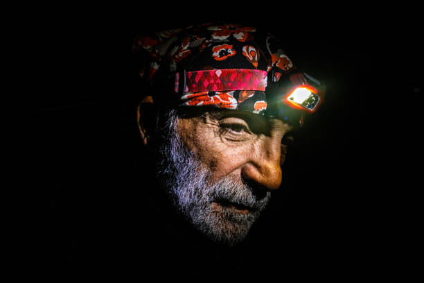 60 years old man with white beard with bandana on his head and flashlight on. The background is black.Close-up portrait with a tired expression