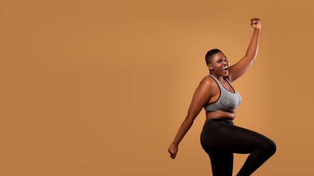 Portrait of emotional black woman cheering and dancing Excited black chubby woman in sportswear dancing, emotional happy lady celebrating success and victory, jumping, cheering and raising clenched fist up, brown studio background, banner, free copy space huge black woman pictures stock pictures, royalty-free photos & images