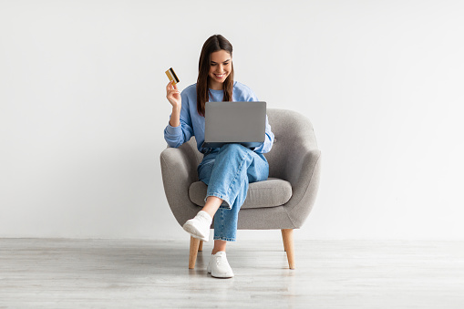Compras en línea, comercio electrónico, banca remota. Mujer joven sentada en sillón con computadora portátil y tarjeta de crédito, comprando en la web photo