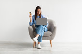 Online shopping, e-commerce, remote banking. Young woman sitting in armchair with laptop and credit card, buying on web